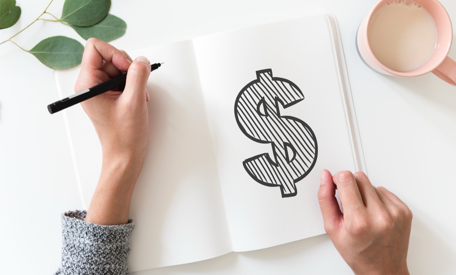 Person drawing dollar sign on paper with black pen, coffee cup nearby